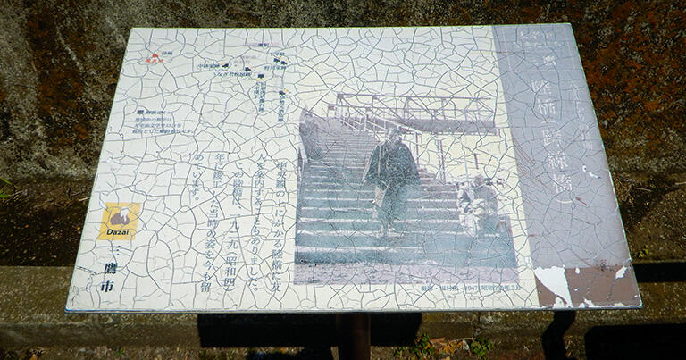 三鷹跨線人道橋のたもとに設置された案内板には、橋を渡る太宰の写真がプリントされている。ヒビで見えにくいものの、まだ補強の斜材が取り付けられておらず、トラスが逆ハの字になっているのが分かる